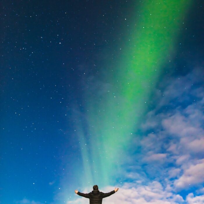 man standing on rock under green aurora borealis and white clouds