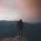 man wearing black bubble jacket on rocky mountain hill in front of mountains under white, pink, and blue cloudy skies during daytime