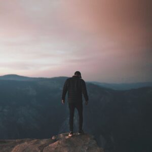 man wearing black bubble jacket on rocky mountain hill in front of mountains under white, pink, and blue cloudy skies during daytime